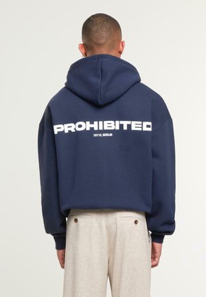 Man wearing navy hoodie with "PROHIBITED 10119, BERLIN" text on back, paired with beige trousers, standing facing away against light background.