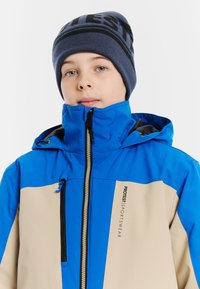 Kolorowa zimowa kurtka z niebieską górną częścią, beżowym dołem, z zamkiem błyskawicznym, wysokim kołnierzem i detalem z logo. Modelka nosi ciemnoniebieską dzianinową czapkę.