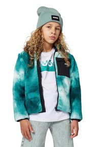 Turquoise fleece jacket with black pocket detail, zip closure, and soft texture. Paired with a white t-shirt and light gray jeans.