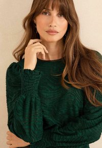 Top verde strutturato con maniche a sbuffo, caratterizzato da un motivo ondulato e dettagli smock alle estremità delle maniche. Tessuto morbido con scollatura rotonda.