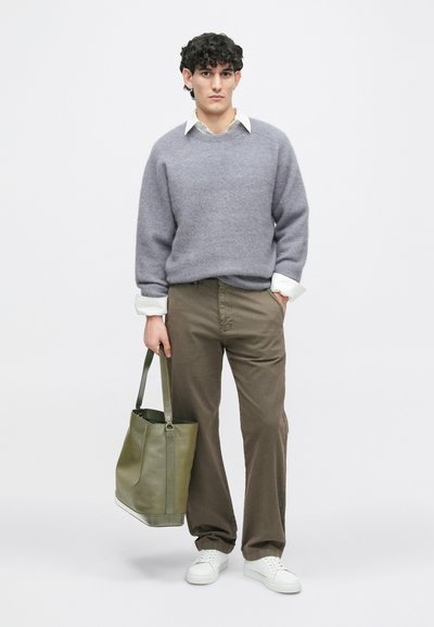 Suéter cinza de lã, camisa branca com colarinho, calças verde-oliva, ténis brancos e uma mala em pele verde. Modelo posicionado ao centro de um fundo neutro.