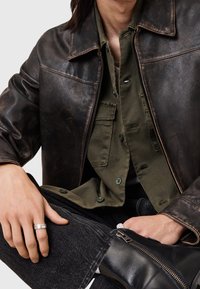 Braune Lederjacke mit glatter Oberfläche, darüber ein olivgrünes Hemd mit Knöpfen. Dunkle Jeans aus Denim und schwarze Stiefeletten sind sichtbar.