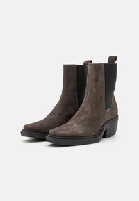 Botas de tobillo de ante marrón con puntera afilada, paneles laterales elásticos y un tacón grueso negro. Textura suave con un acabado mate.