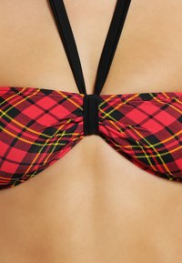 Rood en zwart geruite bikini-top met gele accenten, voorzien van een gladde stof, haltersluitingen en een centrale rimpeling aan de achterkant.