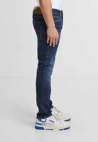 Jeans de mezclilla azul oscuro con un ajuste entallado, que cuentan con una etiqueta de cuero marrón en la cintura. Combinados con zapatillas blancas con acentos azules.