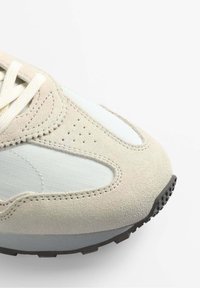 Sneaker en suède beige avec des panneaux en maille gris clair, surface texturée, détails perforés et semelle en caoutchouc noir. Les lacets sont de couleur crème.