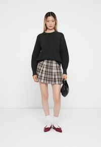 Černý žebrovaný svetr, károvaná mini sukně v hnědých a bílých odstínech, bílé ponožky, červené mokasíny a černá kabelka. Modelka stojí na bílém pozadí.