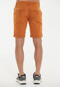 Orange Baumwoll-Shorts mit umgeschlagenem Saum, ausgestattet mit zwei Gesäßtaschen mit Knöpfen, getragen mit grauen Sportschuhen.