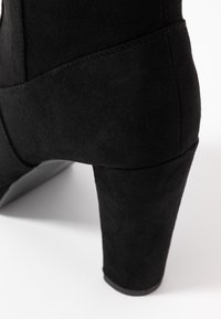 Stivale in suede nero con tacco a blocco, dalla texture liscia e punta leggermente appuntita. Presenta un dettaglio di cucitura sul lato per un design aggiuntivo.