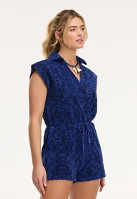 Mėlynas raštuotas rompers turi trumpas rankoves, color ir juosmens juostą. Dekoratyvūs akcentai ir sudėtingi dizainai sustiprina jo tekstūrą.