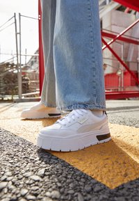 Sneaker platform bianche con una texture liscia, accenti a contrasto e design con lacci. Indossati con jeans a gamba larga azzurri su un attraversamento pedonale.