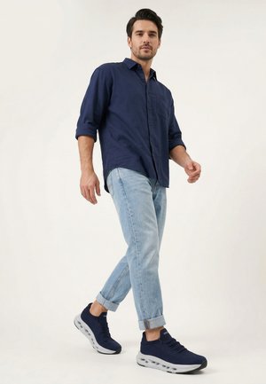 Muž chodí, má na sobě tmavě modrou košili, světle modré ohrnuté džíny a tmavě modré tenisky s bílými podrážkami na jednoduchém pozadí.