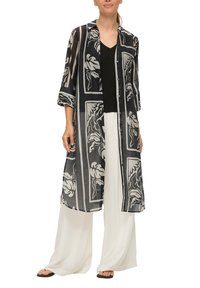 Schwarz-weiß gemusterte lange Bluse mit Kragen, durchgehender Knopfleiste und Dreiviertelärmeln, über einem schwarzen Oberteil und weißen weit geschnittenen Hosen getragen.