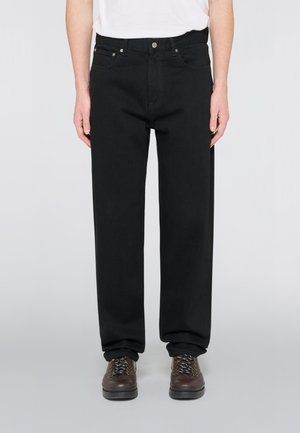 Persona che indossa pantaloni neri dritti, camicia bianca e scarpe marroni con lacci, in piedi su uno sfondo neutro.