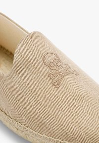 Beige espadrille med fiskebensmønster, elastisk åbning og broderet dødningehoved og korsede knogler detalje. Rebsål synlig ved kanten.