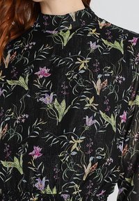 Schwarzes Blumenkleid mit hohem Ausschnitt, leichtem Textil und bunten Blumendrucken in Grün-, Lila- und Pinktönen.