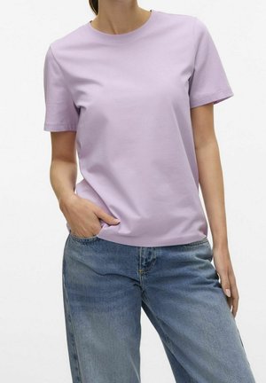 Femme portant un t-shirt uni violet clair à manches courtes et un jean bleu, avec une main dans la poche, sur fond blanc.