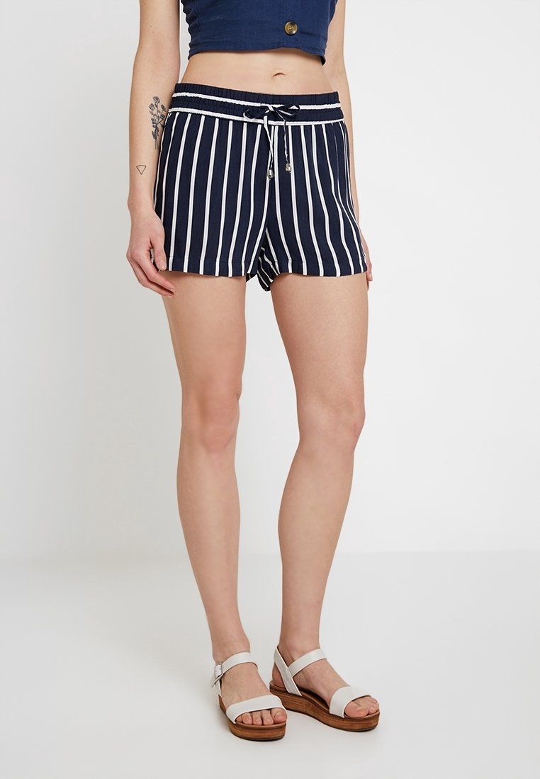 Pantaloni corti a righe blu navy con vita elasticizzata e lacci, caratterizzati da righe bianche e un fit rilassato, abbinati a sandali bianchi.
