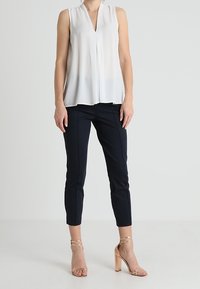 Blusa bianca senza maniche con scollo a V, abbinata a pantaloni attillati blu navy e sandali con tacco a strappo color nude. Tessuto liscio e leggero.