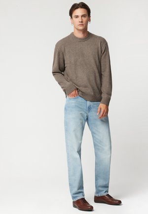 Brązowy sweter z dzianiny z okrągłym dekoltem, zestawiony z jasnoniebieskimi dżinsami i brązowymi skórzanymi butami. Model stoi z rękami w kieszeniach.