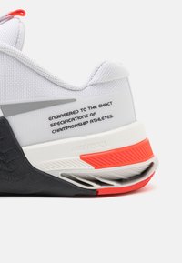 Scarpa sportiva bianca con tomaia in rete testurizzata, suola in gomma nera e dettagli rossi. Presenta testo stampato sul tallone. Marchio Nike React visibile.