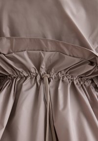 Tissu beige avec une taille élastiquée et un cordon de serrage ajustable doté d'un toggle en plastique pour le resserrer ou le desserrer.