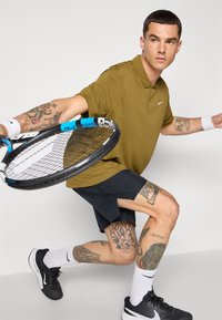 Grønn polo skjorte, svarte shorts, svarte joggesko, hvite sokker. Holder en tennisracket med blå detaljer. Synlige tatoveringer på armer og ben.