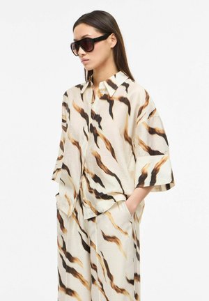 Nainen, joka pukeutuu ylisuurteen kermaan ja samanlaisiin leveälahkeisiin housuihin, joissa on ruskeita ja mustia abstrakteja raita kuvioita, yhdistettynä suuriin mustiin aurinkolaseihin.