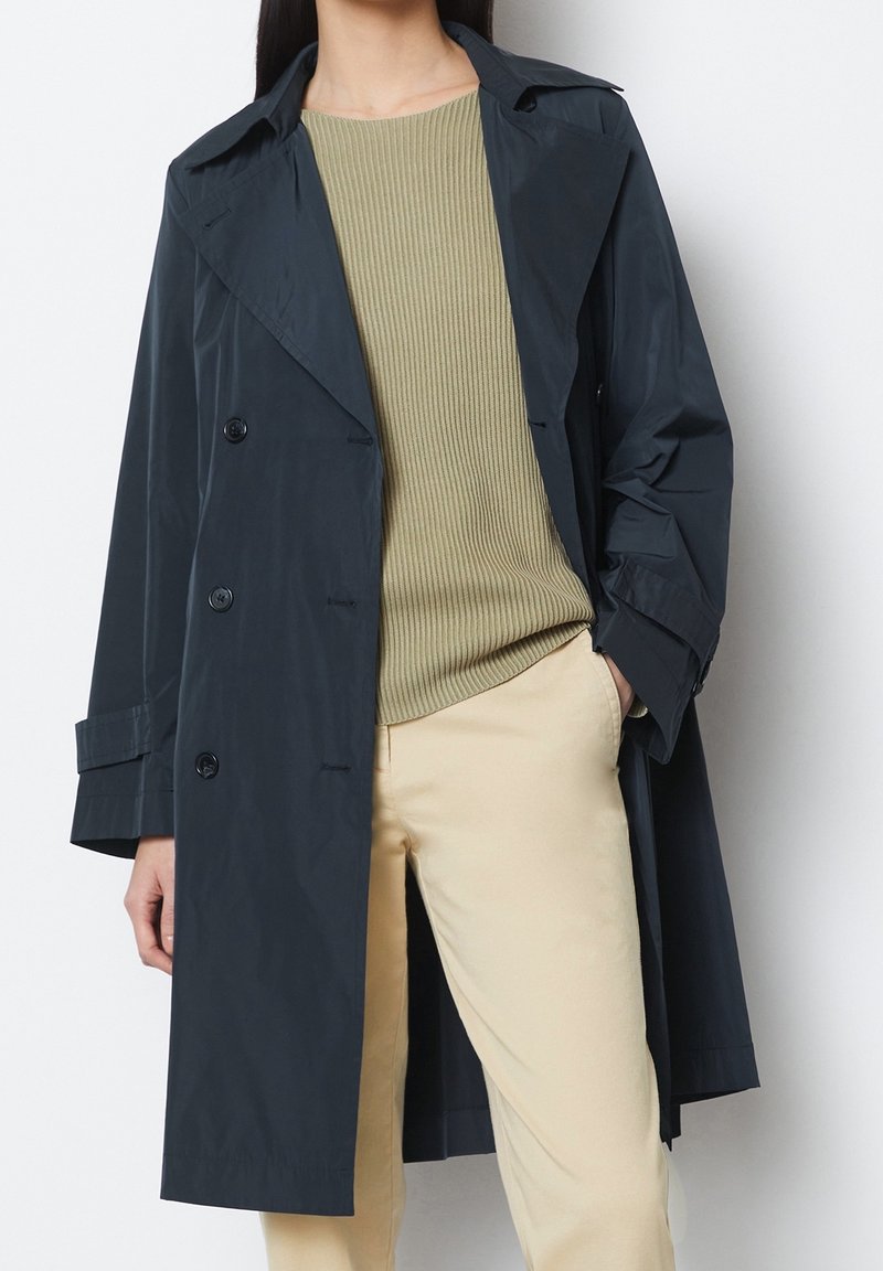 Personne portant un trench-coat bleu marine foncé par-dessus un pull côtelé vert olive clair et un pantalon beige, debout avec une main dans la poche.