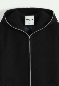 Veste à capuche noire en tissu doux, avec une fermeture éclair argentée et des poches avant. L'étiquette indique la marque FRILIVIN et la taille M.