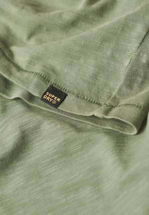 Tissu vert avec un tissage texturé, présentant une petite étiquette noire sur laquelle est inscrit "SUPER DRY" en lettres dorées, partiellement visible sur le bord.