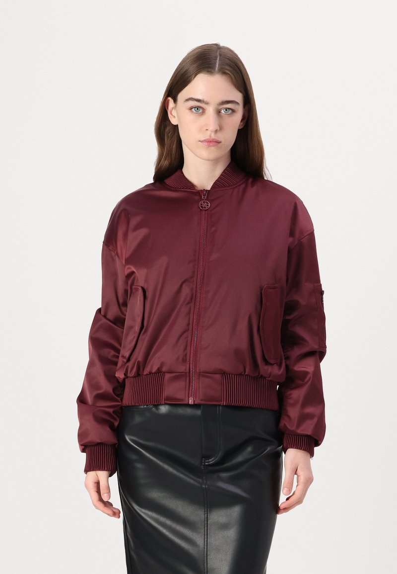 Giacca bomber in satin borgogna con polsini e orlo a coste, dotata di tasche laterali e chiusura frontale con zip. Indossata sopra una gonna in pelle nera.