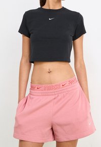 Černé zkrácené tričko s krátkými rukávy a bílým logem Nike, kombinované s růžovými šortkami Nike s kontrastním pasem a bočními kapsami.