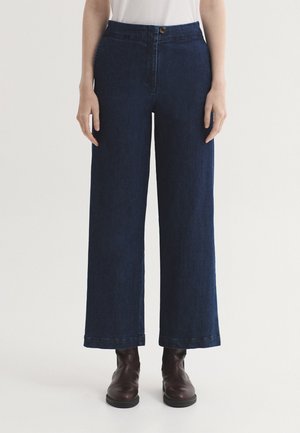 Personne portant un jean bleu foncé à jambes larges avec fermeture par bouton et zip, associé à des bottines marron foncé, debout sur un fond uni.