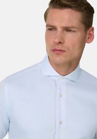 Mann mit kurzem braunem Haar, der ein hellblaues, strukturiertes Hemd mit geschlossenem Kragen trägt und seitlich vor einem schlichten Hintergrund schaut.