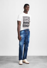 Vit t-shirt med kort ärm och svart grafiskt tryck, ihop med blå slitna jeans och krämfärgade sneakers. Enkel design, avslappnad passform.