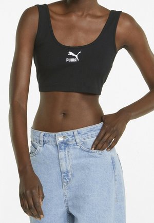 Femme portant un crop top noir Puma et un jean taille haute bleu clair, une main dans la poche du jean et l'autre détendue le long du corps.