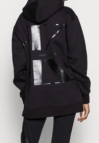 Czarna bluza z kapturem z srebrnym, odblaskowym wzorem na plecach, o luźnym kroju i kapturze. Wykonana z miękkiej, teksturowanej tkaniny.