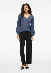 Suéter de punto azul con un escote en V con bordes ondulados y mangas globo texturizadas, combinado con pantalones rectos negros y zapatos de punta negra.