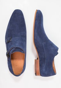 Chaussures en daim bleu marine avec un bout pointu, deux accents de boucles et un talon en bois. Le design présente une texture lisse et une silhouette incurvée.
