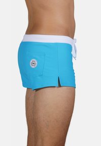 Shorts de bain bleu clair avec une taille blanche, fente latérale et poche arrière avec un patch logo rond. Fabriqué à partir de matériau à séchage rapide.