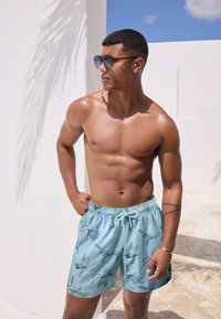 Pantaloni scurți de înot albastru deschis cu un model floral, având talie cu șnur. Materialul pare neted și ușor. Fără detalii suplimentare.