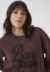 Bordeauxfarbener Sweatshirt mit schwarzem gesticktem Text "Bonjour." Verfügt über einen Rundhalsausschnitt und eine lockere Passform; der Stoff wirkt weich mit einer glatten Textur.