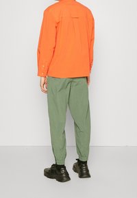 Pomarańczowa koszula na guziki z plisą z tyłu, zestawiona z zielonymi, zwężanymi spodniami. Czarne trampki dopełniają strój. Jednolite tło.