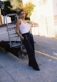 Une femme portant un débardeur blanc et un pantalon marine s'appuie contre un bateau nommé « Freya » sur un quai baigné de soleil avec des plantes en arrière-plan.