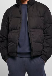 Chaqueta negra tipo puffer con un alto cuello, paneles acolchados, cierre frontal con cremallera y ajuste ceñido. Llevada sobre un sudadera azul marino.