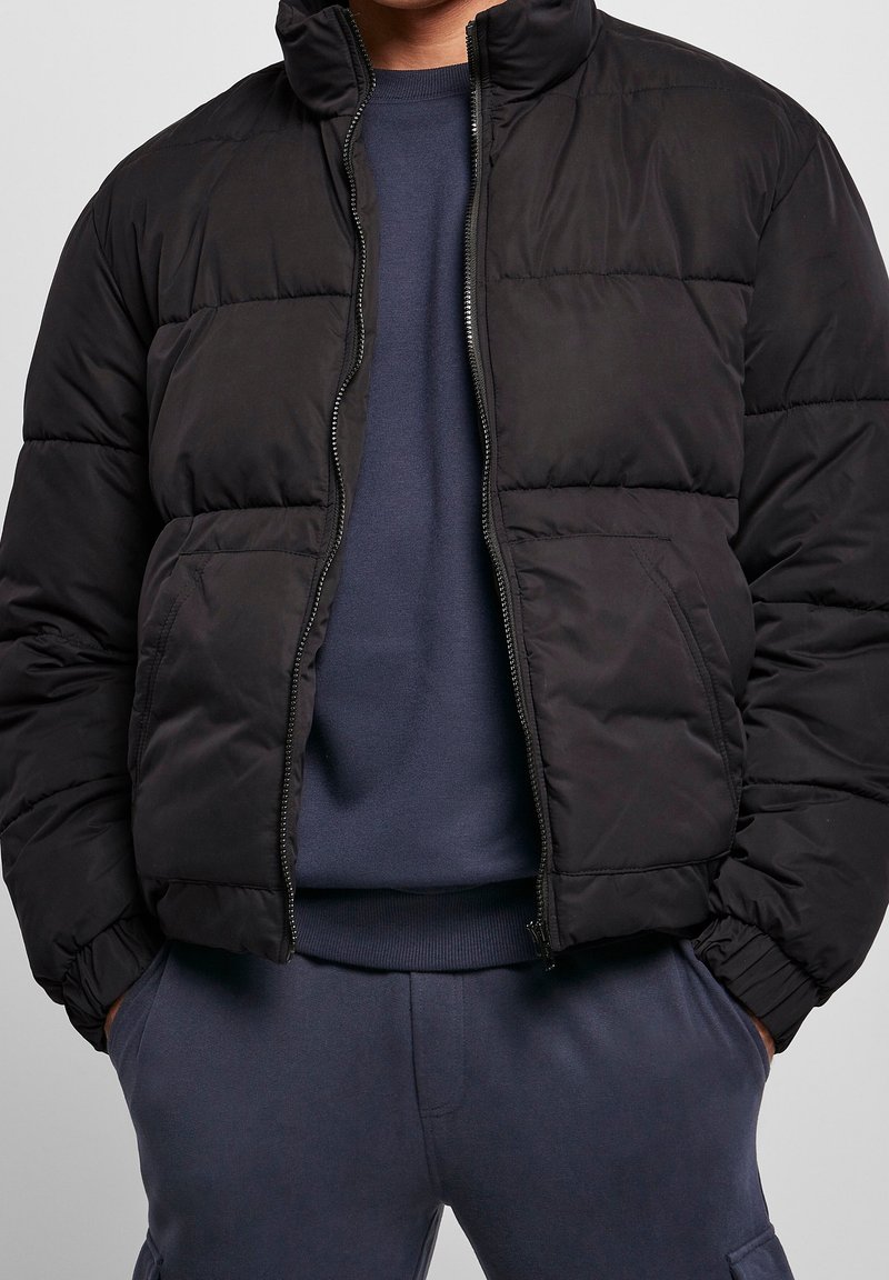 Giubbotto a vento nero con colletto alto, pannelli trapuntati, chiusura con zip e vestibilità aderente. Indossato sopra un felpa blu navy.