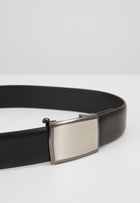 Sort læderbælte med en rektangulær sølvmetalspænde, der har en børstet overflade og en dekorativ accent på den ene side.
