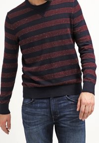 Mann trägt einen langärmligen Pullover mit bordeauxroten und marineblauen Streifen und dunkle blaue Jeans, steht vor einem weißen Hintergrund.