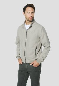 Chaqueta gris claro con cremallera, hecha de tela suave, con cuello alto, puños acanalados y bolsillos laterales. Se lleva sobre una camiseta blanca.